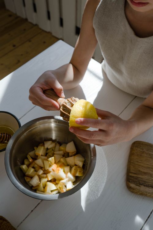 Bir çocuk yetişkinin yardımıyla meyve hazırlarken limonu doğranmış meyvelerin bulunduğu kaba sıkıyor.